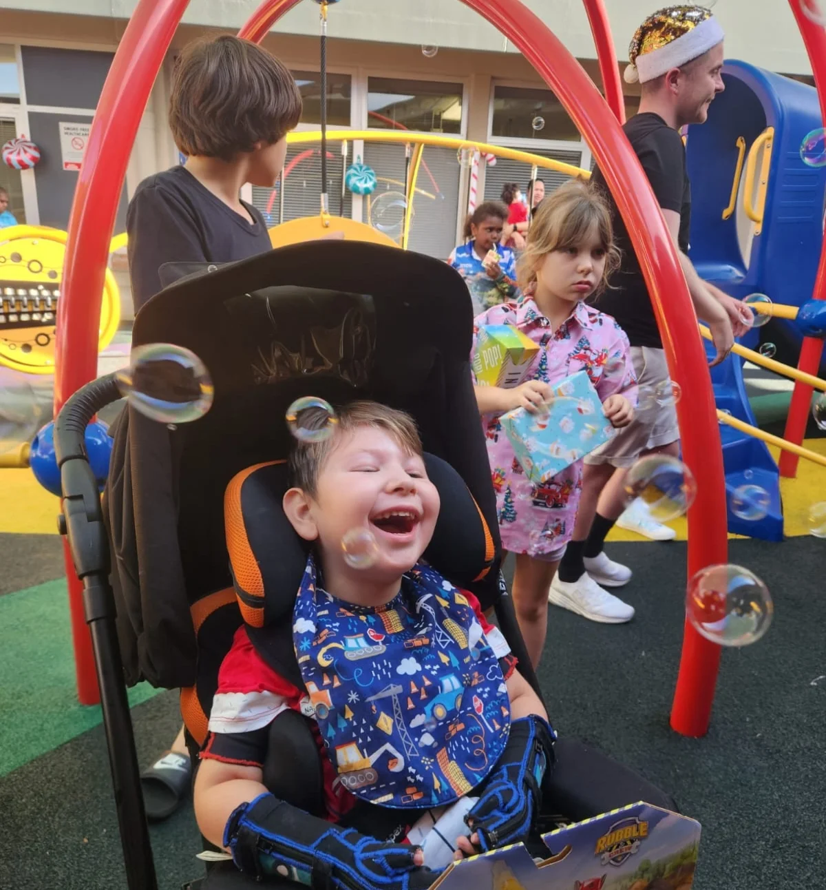 Child in a wheelchair laughs at bubbles, surrounded by other children in a playground.