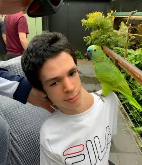 A green parrot sits on the shoulder of a person with short dark hair wearing a white FILA shirt. They are outdoors, with greenery and another person in the background.