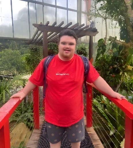A person stands on a red wooden bridge in a greenhouse, wearing a red shirt and shorts, with a backpack. Lush greenery surrounds the area.