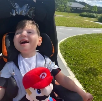 A child in a stroller is smiling while holding a Mario plush toy. They are outdoors on a sunny day, with a path and grassy area in the background.