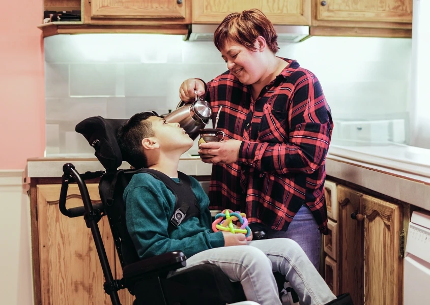 A person in a red flannel shirt is pouring a drink for a child sitting in a wheelchair, holding a colorful toy, in a kitchen setting.