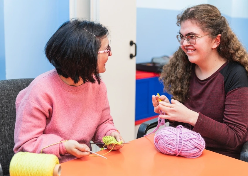 Two people sitting at a table, smiling and engaging in knitting, surrounded by colorful yarn.