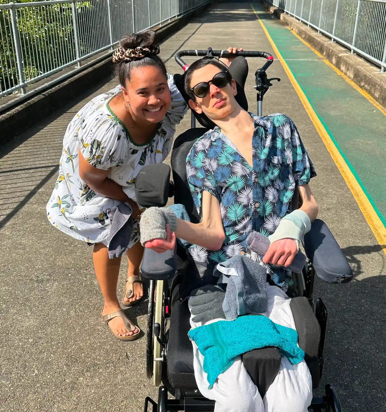 Two individuals on a bridge. One stands and smiles, while the other, in sunglasses, sits in a wheelchair. Clothing items are on the wheelchair.