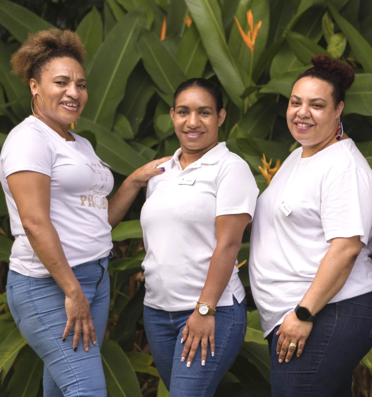 Three people in white shirts and jeans stand together outdoors in front of green foliage.