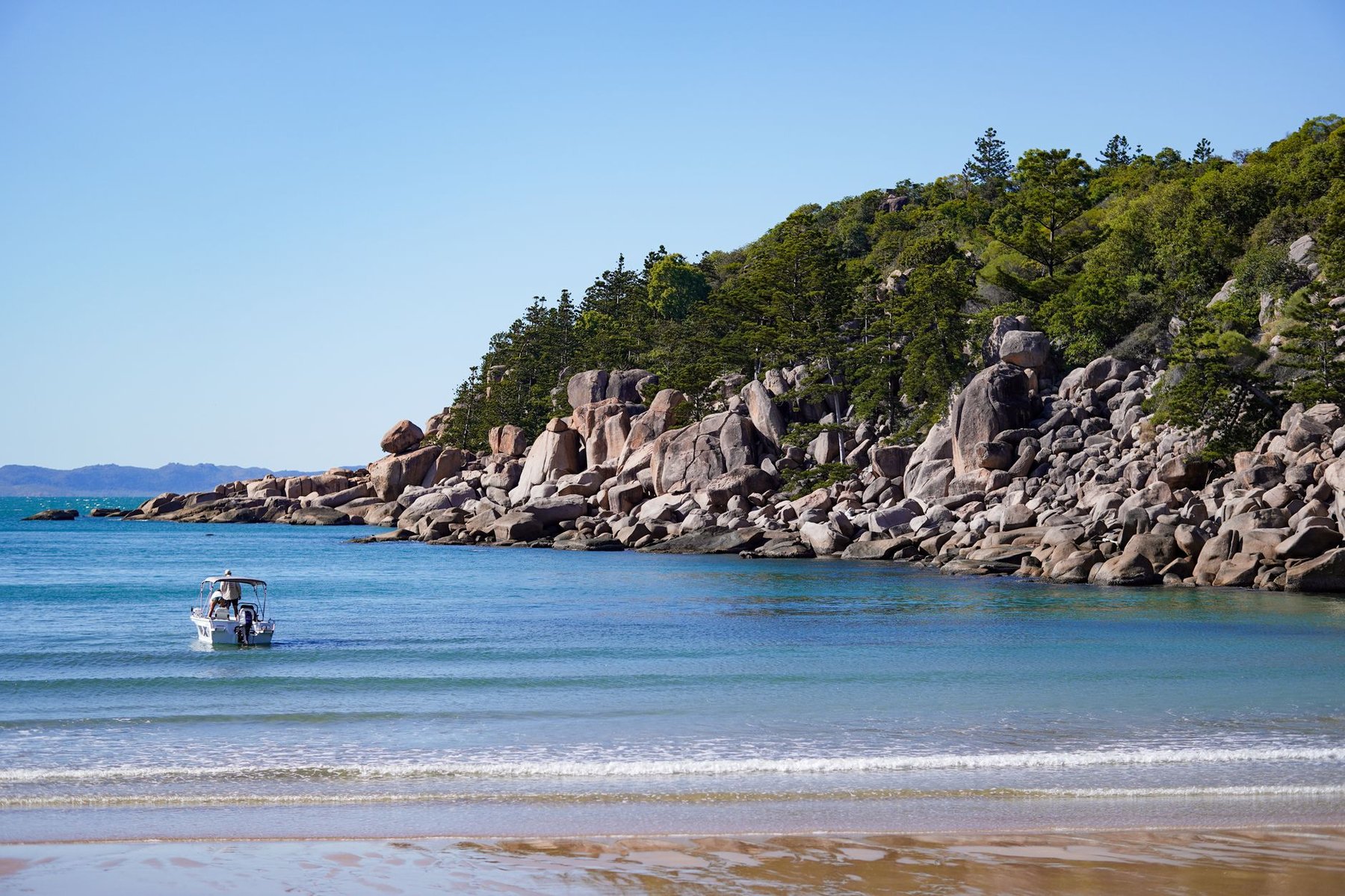 A small boat approaches a tranquil turquoise bay lined with granite boulders and tropical trees, a classic accessible day-trip destination near Townsville, Queensland.