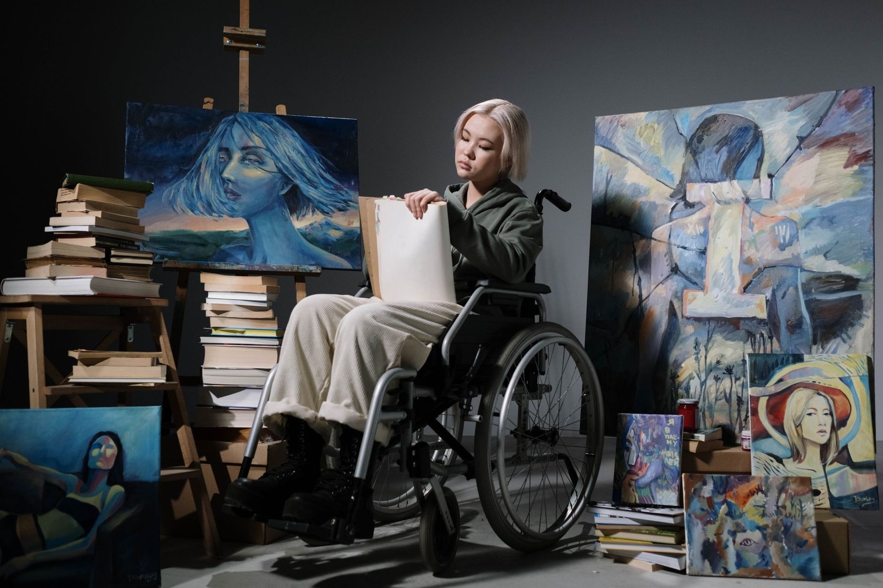 A young artist in a wheelchair surrounded by her colourful paintings in a creative studio, representing inclusive arts programs for people with disability