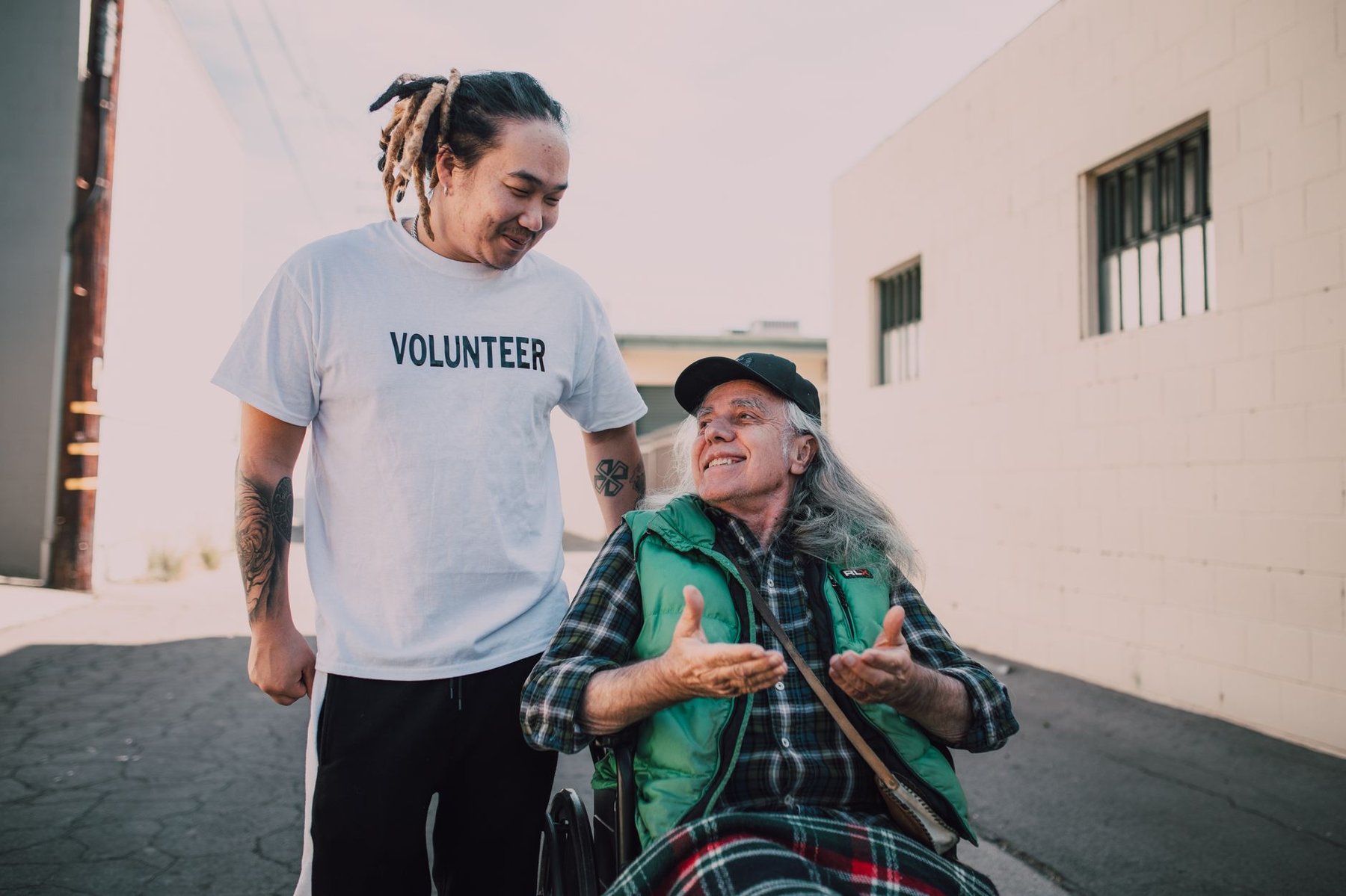 Smiling volunteer wearing a "Volunteer" t-shirt warmly chatting with an older man in a wheelchair outdoors, representing community disability support programs.