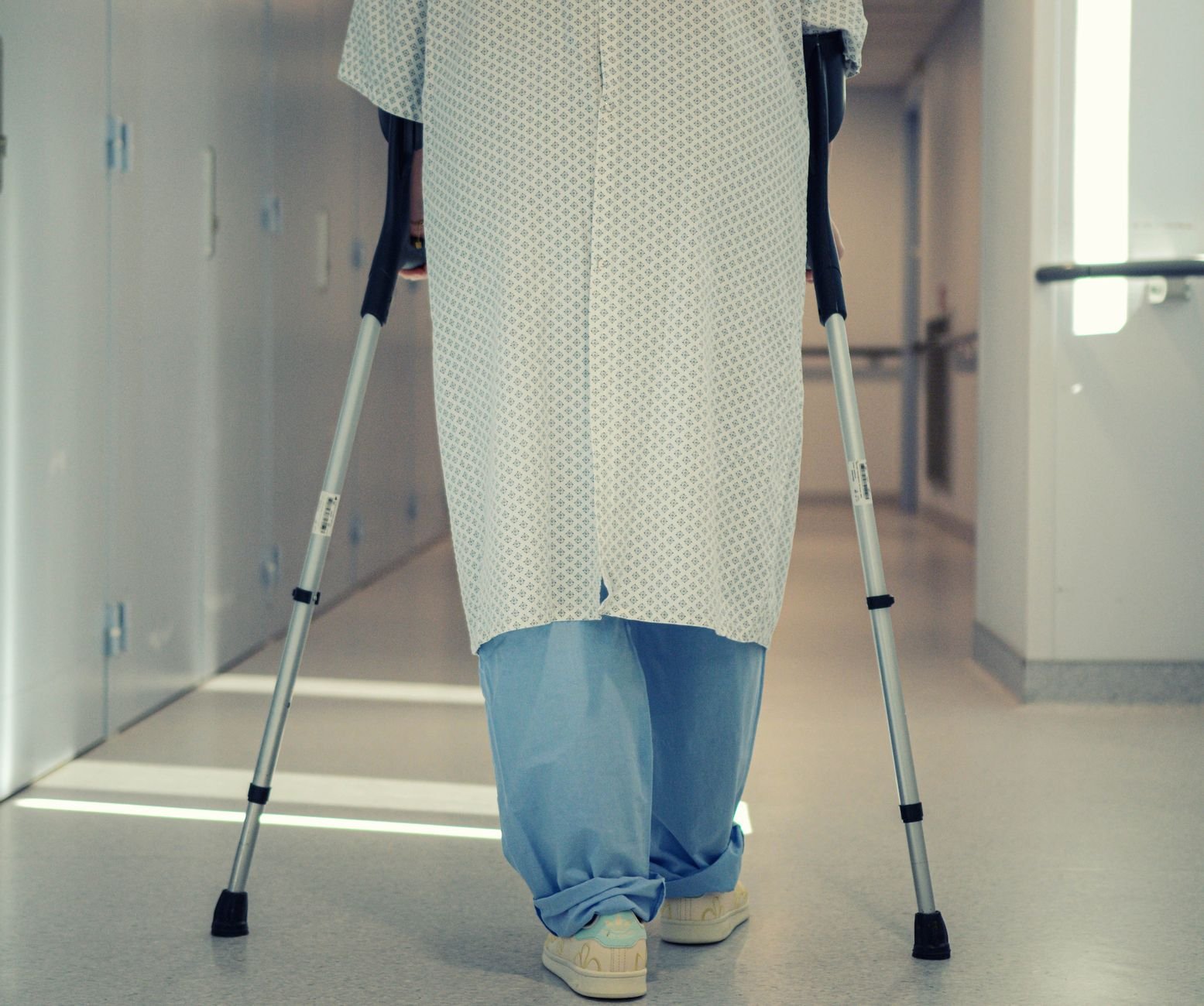 A patient in a hospital gown walks down a corridor using crutches, symbolizing recovery and the need for support after hospital discharge