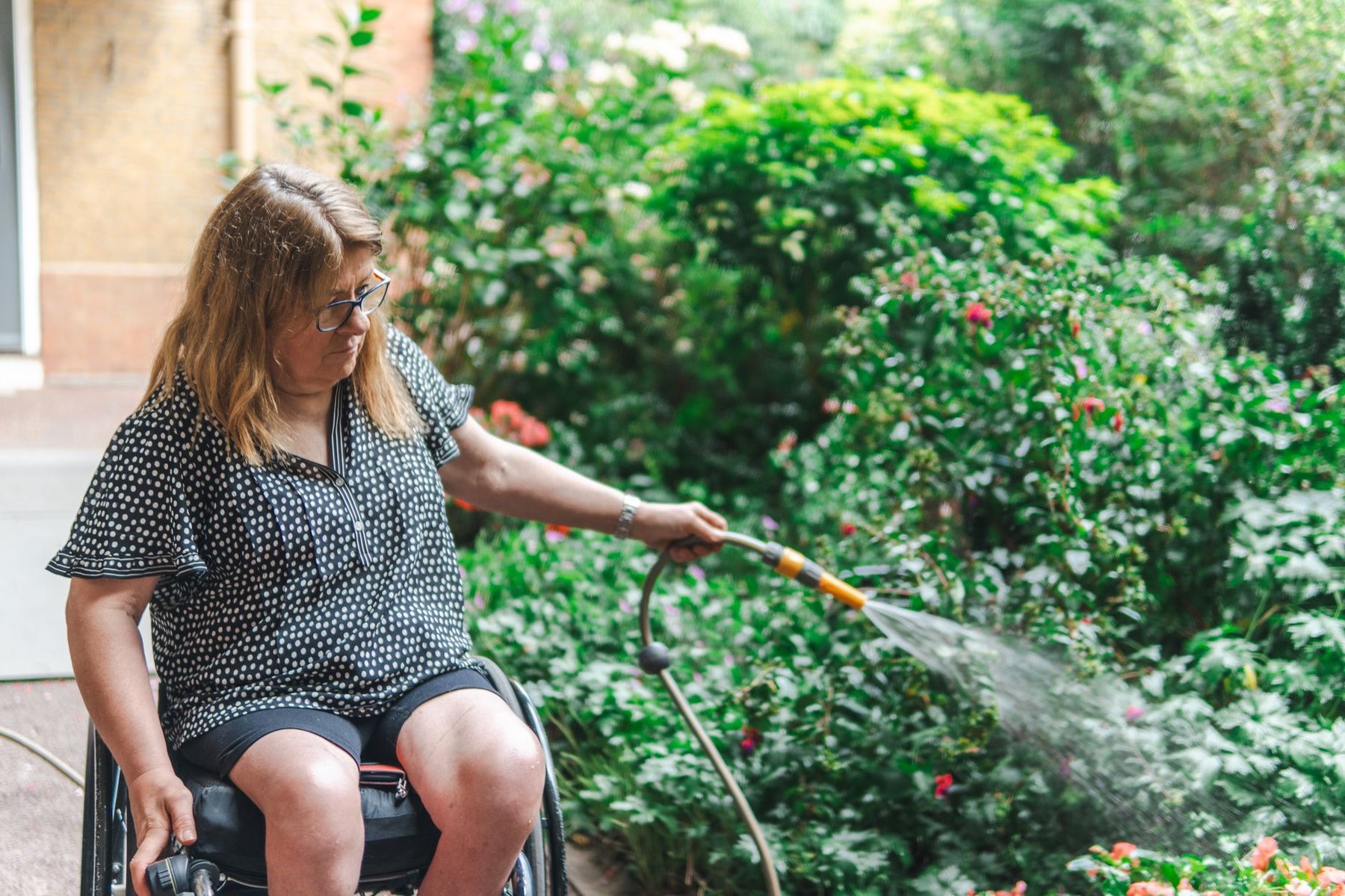 Woman in a wheelchair confidently watering flowers in a lush garden, demonstrating independence built through NDIS capacity building support