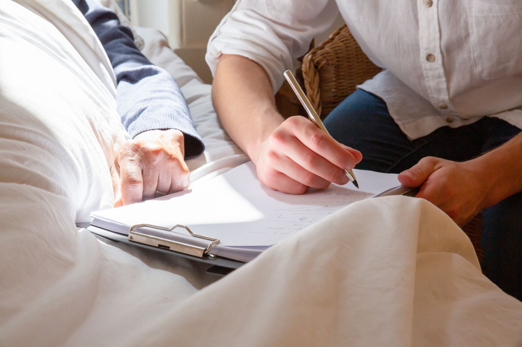 Support worker writing care notes on a clipboard beside a client resting in bed, lit by warm sunlight
