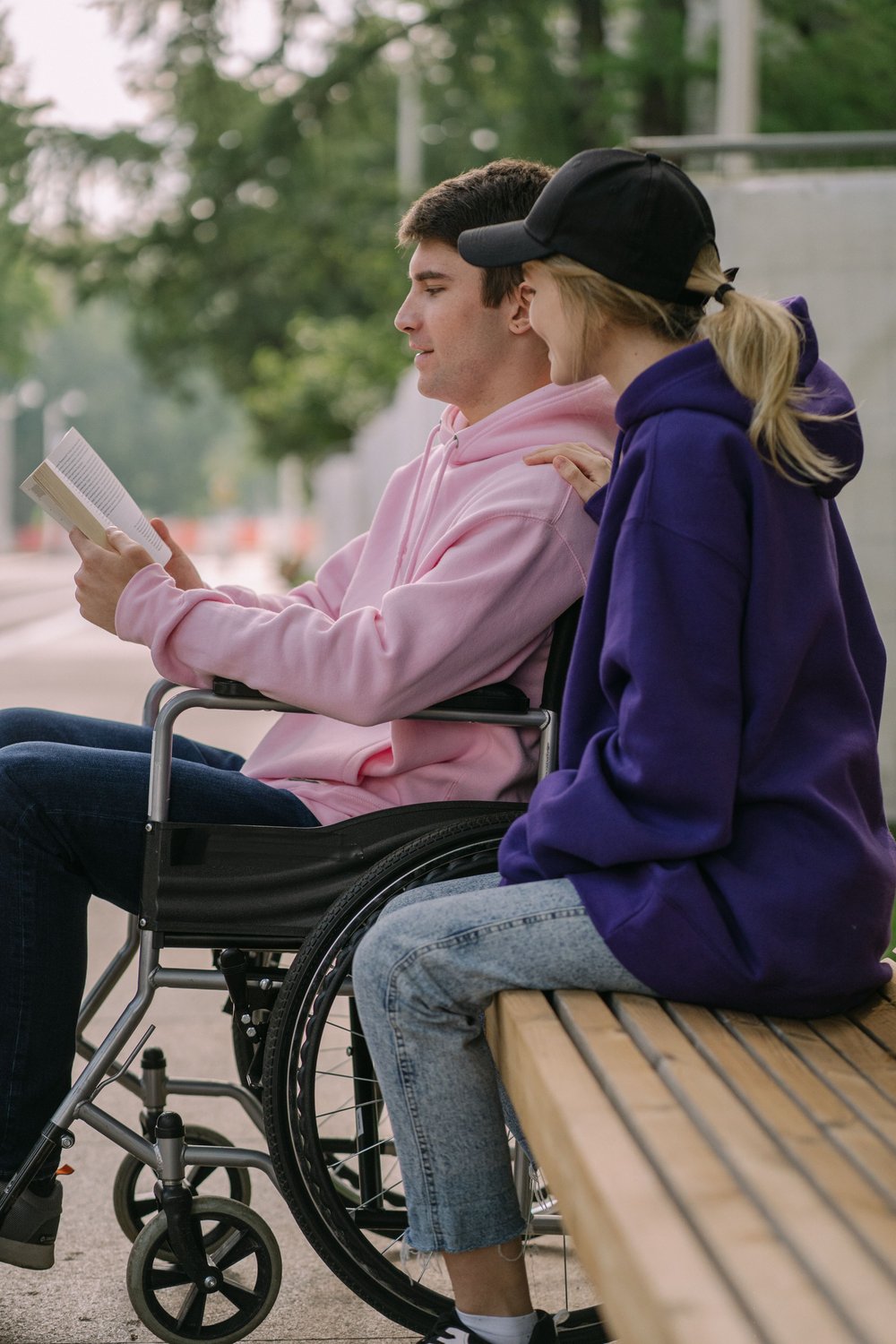 A young man in a wheelchair reading a book outdoors with a companion beside him on a sunny day, reflecting the personal side of disability support services.