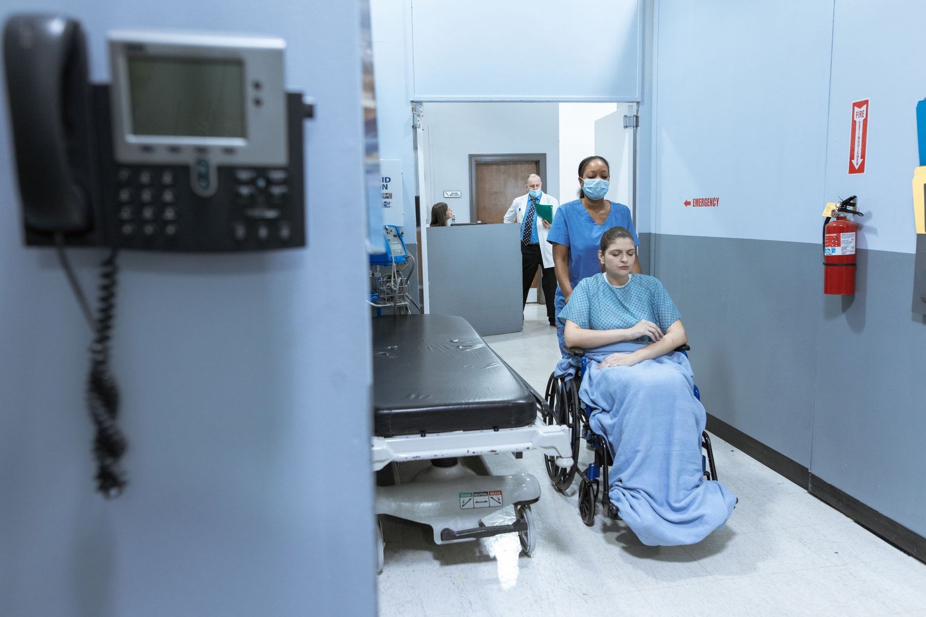 A nurse in scrubs and mask carefully wheels a patient wrapped in a blanket through a hospital corridor, representing the transition of complex care needs at discharge.