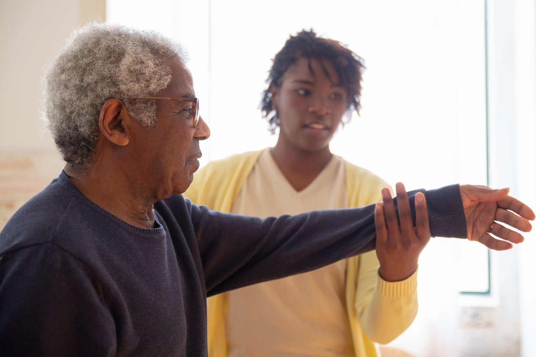 Older man doing arm rehabilitation exercises at home with a caregiver's help while recovering after a stroke