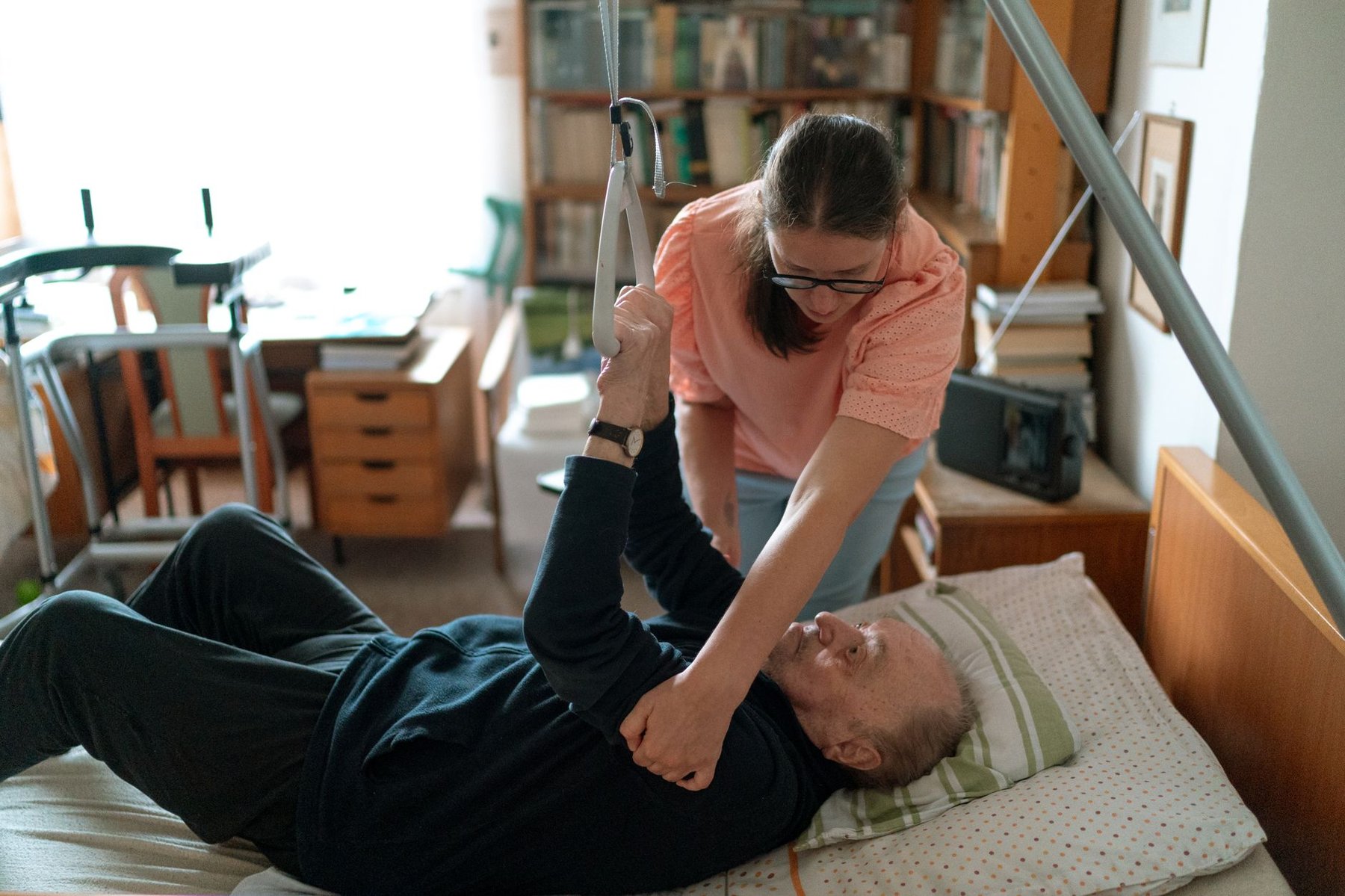 Carer helping a man in a home care bed use a trapeze bar to reposition himself, with a walker nearby, during recovery at home after a brain injury
