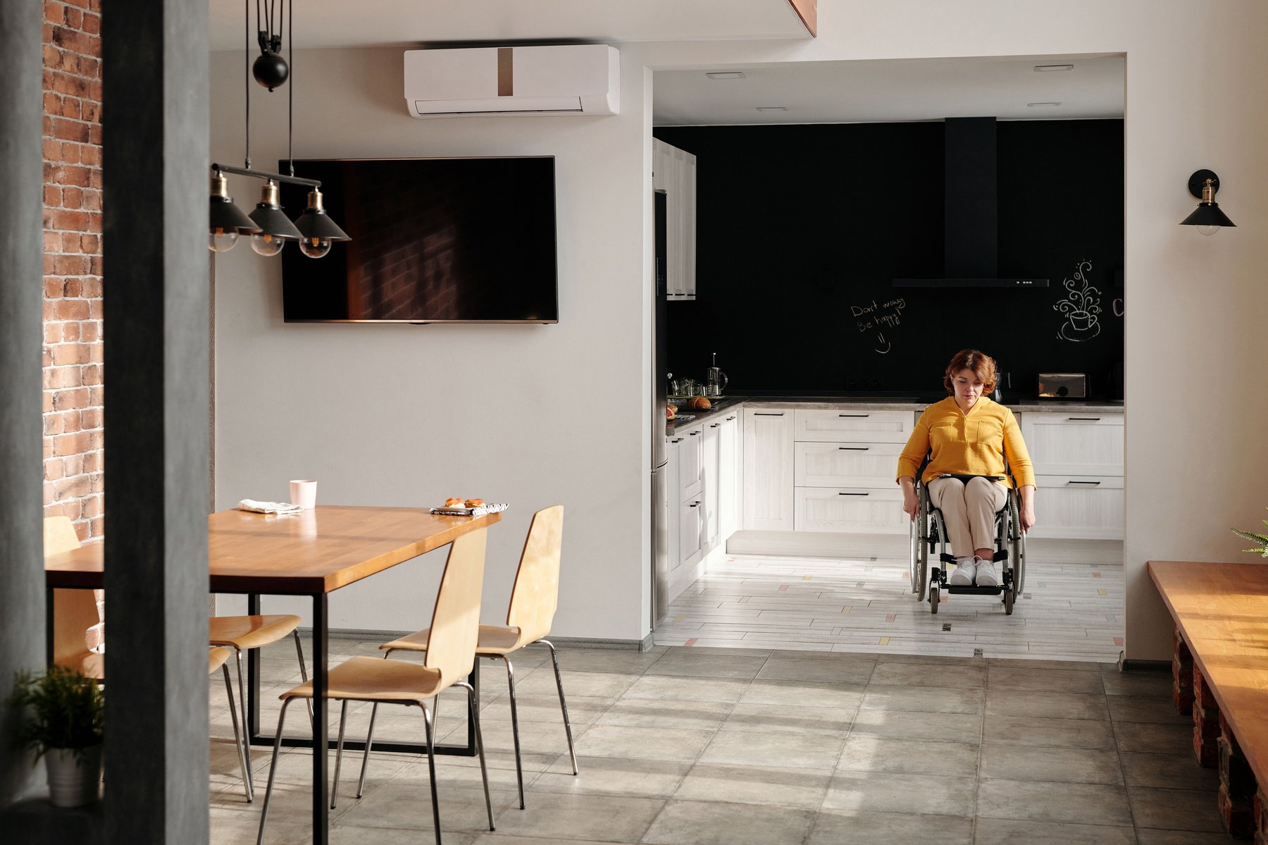 Woman using a wheelchair moving through an accessible, modern kitchen and living space, representing supported independent living