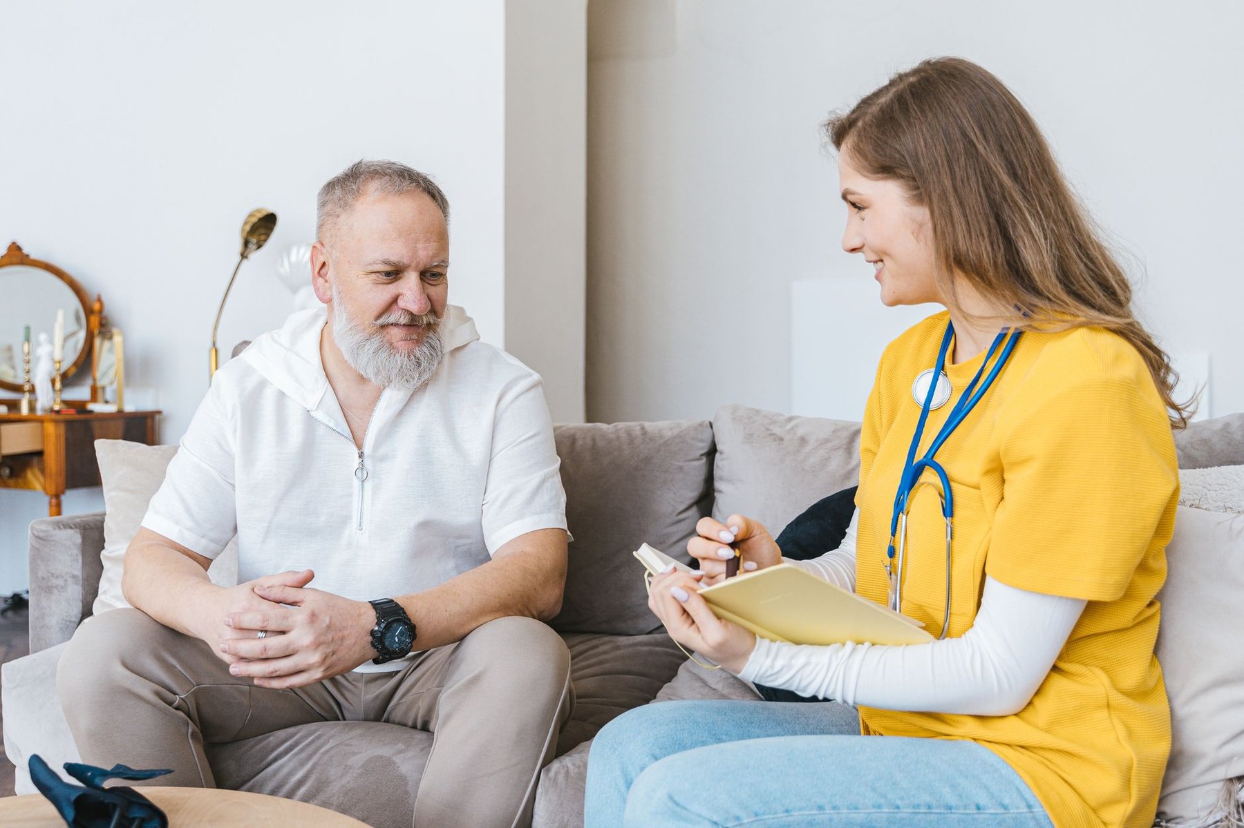 Home care nurse with stethoscope taking notes during a care handover conversation with an older man in his living room