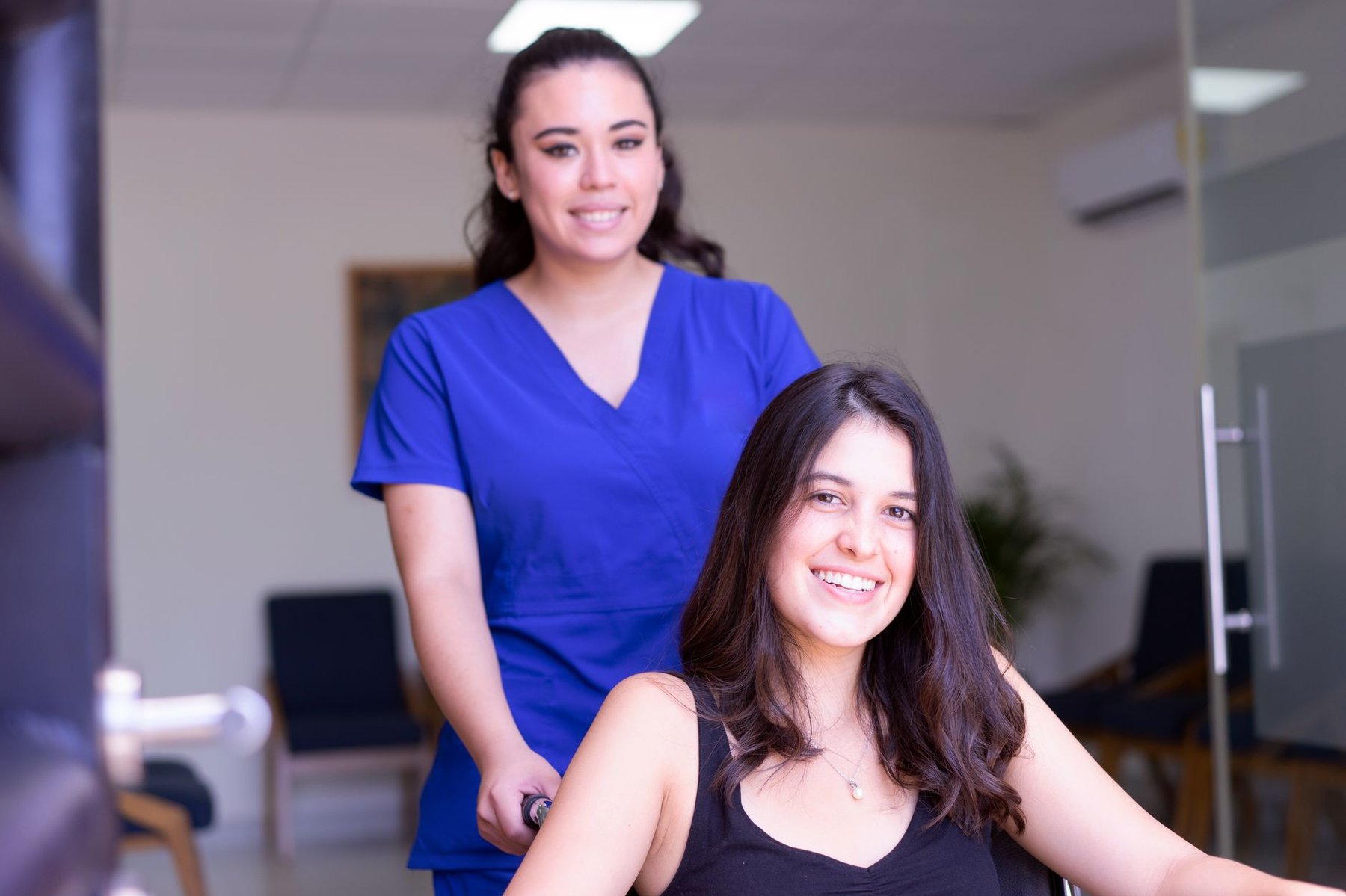 Professional support worker in scrubs assisting a smiling client in a wheelchair in a bright clinical setting