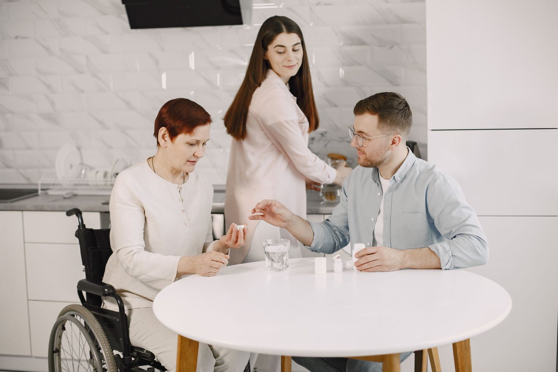 Support worker assisting a wheelchair user with medication at a kitchen table while another person looks on