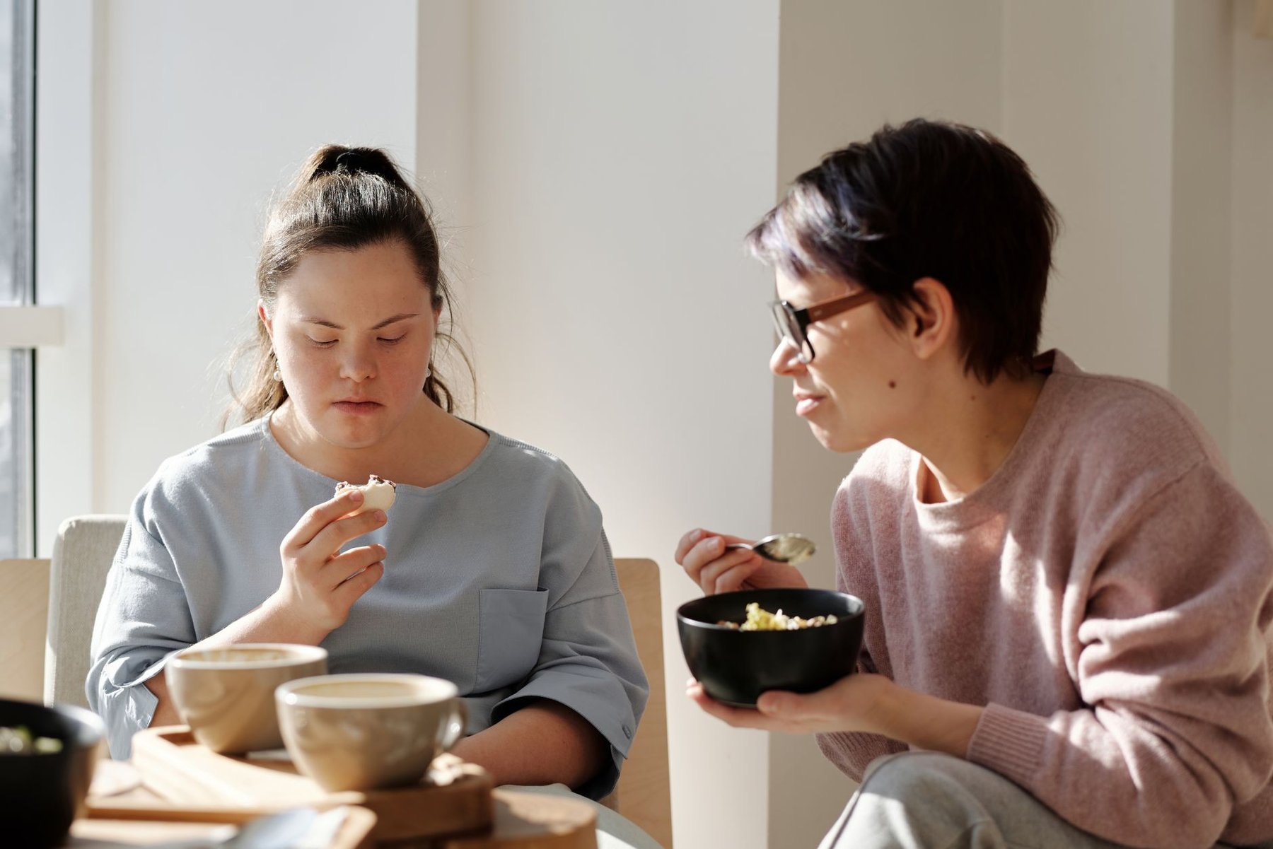 A support worker sharing a meal one-on-one with a young woman with Down syndrome in a sunlit home, showing 1:1 disability support