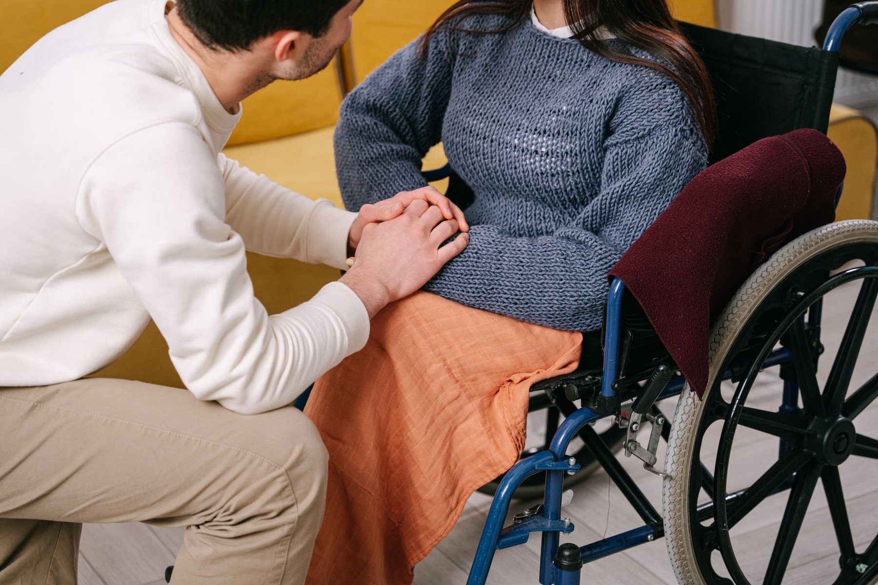 Support worker kneeling beside a woman in a wheelchair and holding her hands, showing reassuring NDIS support for people living with progressive conditions