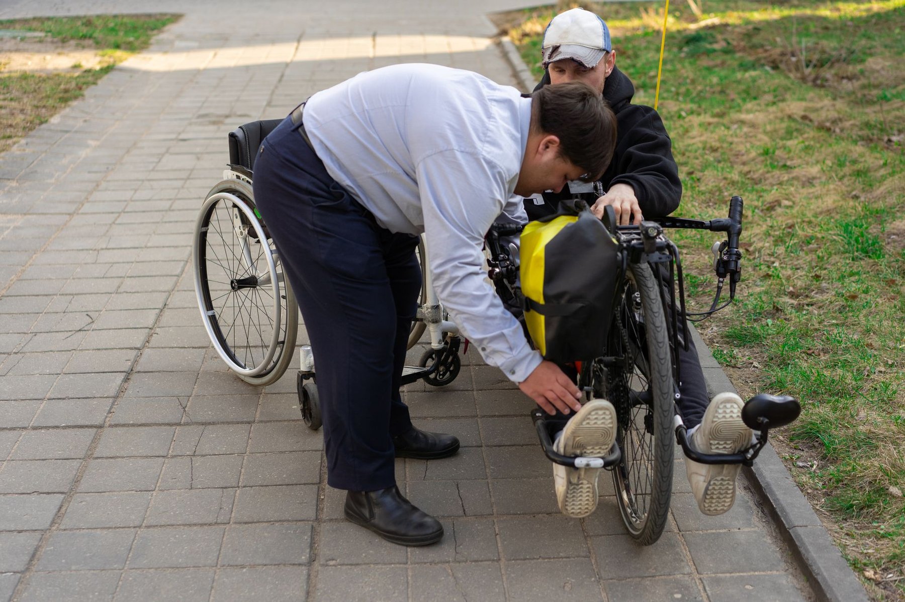 Support worker kneeling to adjust the footrest of a man's wheelchair with a power attachment on an outdoor path, showing hands-on disability support